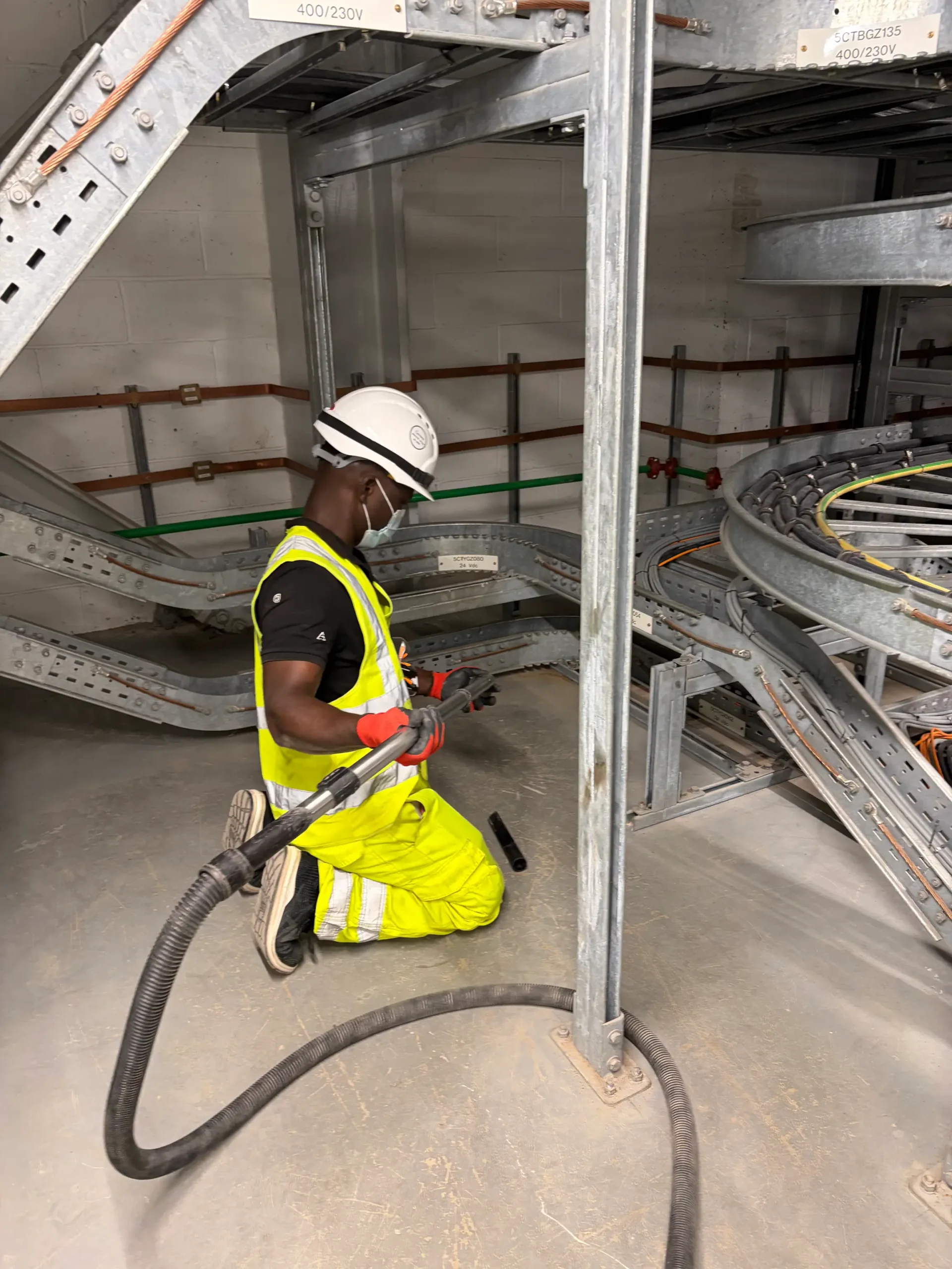 NDH Cleaning Ltd operative in hard hat, hi-vis and face mask vacuuming dust from beneath electrical cable containment in a switchroom
